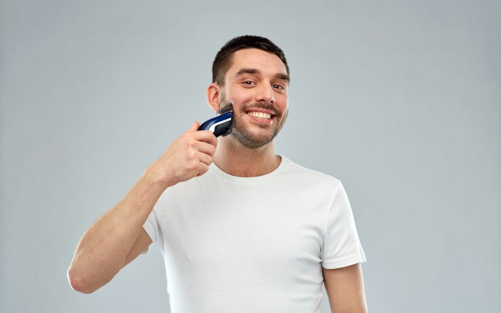beauty, grooming and people concept - smiling young man shaving beard with trimmer or electric shaver over gray background